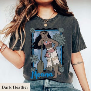 May include: Dark heather grey t-shirt with a graphic of Moana, framed in blue, and the word "Moana" in blue script. The design includes a paddle and floral elements. The shirt's colour is dark heather.