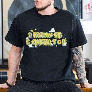 May include: A black short-sleeved t-shirt featuring a graphic print with the text I KNEW IT I KNEW YOU in yellow letters with blue outlines. The design includes small white clouds and yellow stars, with a red star inside the letter O.
