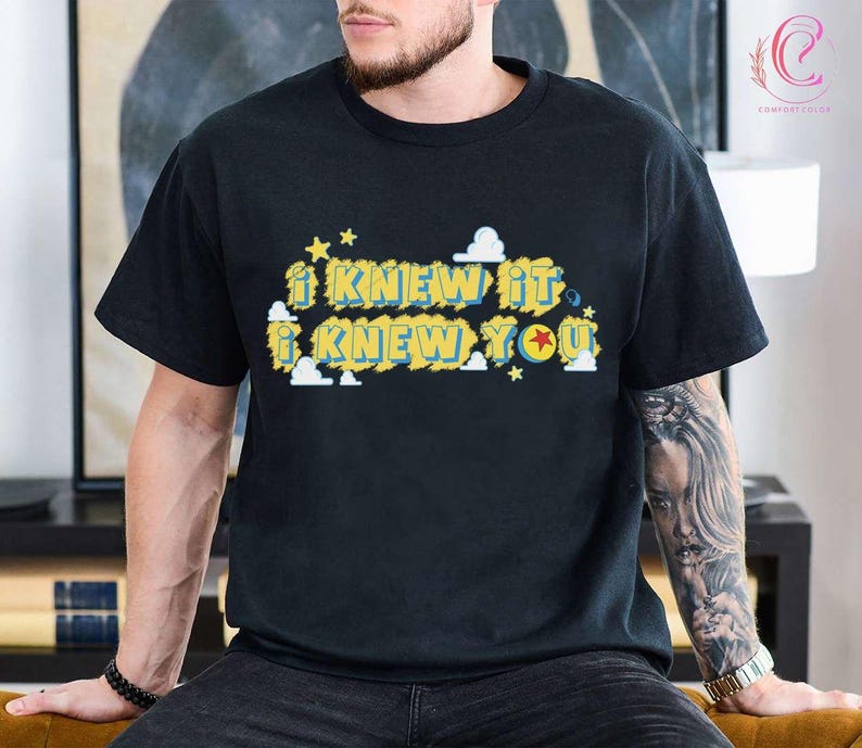May include: A black short-sleeved t-shirt featuring a graphic print with the text I KNEW IT I KNEW YOU in yellow letters with blue outlines. The design includes small white clouds and yellow stars, with a red star inside the letter O.