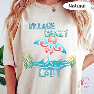K&ouml;nnte beinhalten: Cremefarbenes T-Shirt mit dem Schriftzug "VILLAGE CRAZY LADY" in Blau und Gr&uuml;n. Ein stilisiertes Design eines Mantarochens in Blau, Rot und Pink ist auf dem Shirt zentriert, mit Wellen- und Seegras-Akzenten. Das Wort "Natural" ist oben aufgedruckt.