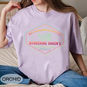 May include: A light purple t-shirt with a graphic that reads "BIG SUMMER BLOWOUT" and "WANDERING OAKEN'S ARENDELLE" in a multi-colored hexagon design. The shirt is a solid color and has a crew neck.