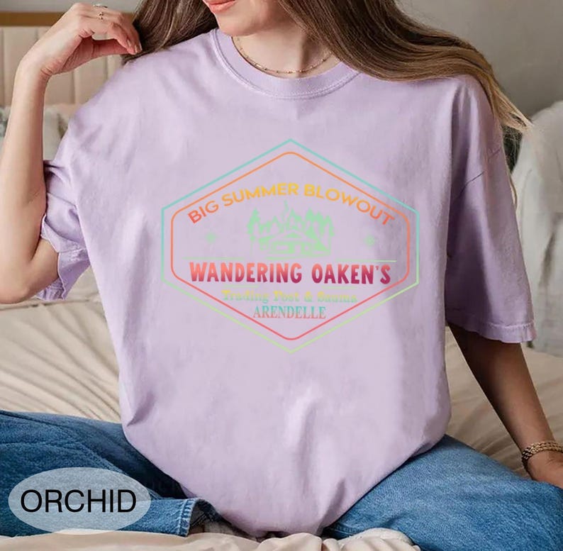 May include: A light purple t-shirt with a graphic that reads "BIG SUMMER BLOWOUT" and "WANDERING OAKEN'S ARENDELLE" in a multi-colored hexagon design. The shirt is a solid color and has a crew neck.