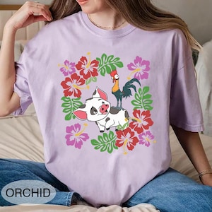 Może przedstawiać: Fioletowa koszulka z nadrukiem świni i koguta otoczonych czerwonymi, r&oacute;żowymi i zielonymi elementami kwiatowymi. Słowo "ORCHID" jest wydrukowane na dole. Koszulka jest wykonana z miękkiego materiału.