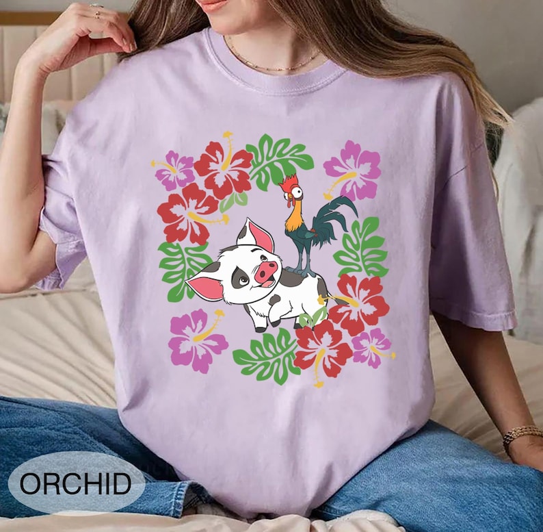 Może przedstawiać: Fioletowa koszulka z nadrukiem świni i koguta otoczonych czerwonymi, r&oacute;żowymi i zielonymi elementami kwiatowymi. Słowo "ORCHID" jest wydrukowane na dole. Koszulka jest wykonana z miękkiego materiału.