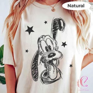 May include: A cream-coloured t-shirt with a black and white sketch of a cartoon dog with its tongue out, surrounded by black stars. The word "Natural" is printed at the top.