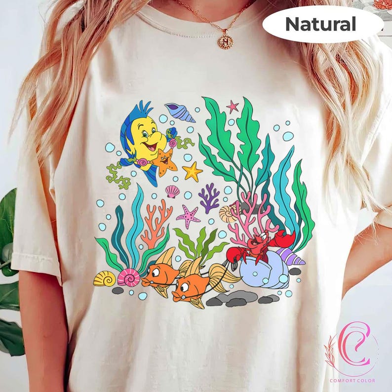 K&ouml;nnte beinhalten: Cremefarbenes T-Shirt mit einer farbenfrohen Unterwasserszene mit Zeichentrickfiguren. Das Design umfasst Fische, eine Krabbe, Seetang und andere Meeresbewohner. Das Wort "Natural" ist oben rechts aufgedruckt.