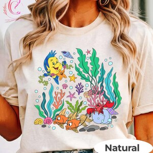 K&ouml;nnte beinhalten: Beiges T-Shirt mit einem farbenfrohen Unterwassermotiv. Die Grafik zeigt Zeichentrickfische, Seetang und verschiedene Meeresbewohner. Ein gelber Fisch, Seesterne und eine rote Krabbe sind abgebildet. Das Wort "Natural" ist unten aufgedruckt.