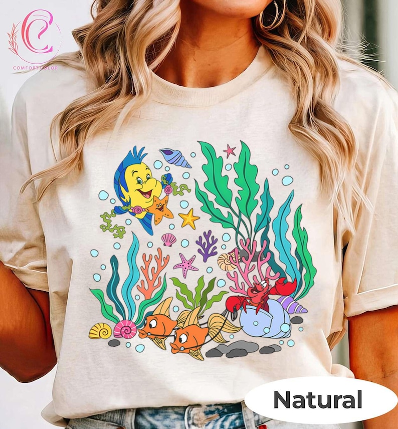 K&ouml;nnte beinhalten: Beiges T-Shirt mit einem farbenfrohen Unterwassermotiv. Die Grafik zeigt Zeichentrickfische, Seetang und verschiedene Meeresbewohner. Ein gelber Fisch, Seesterne und eine rote Krabbe sind abgebildet. Das Wort "Natural" ist unten aufgedruckt.