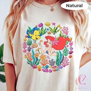 Pu&ograve; includere: T-shirt color crema con una grafica colorata di Ariel e Flounder de La Sirenetta, circondati da coralli, alghe e conchiglie. La parola "Natural" &egrave; stampata in alto a destra. La maglietta &egrave; di Comfort Colors.