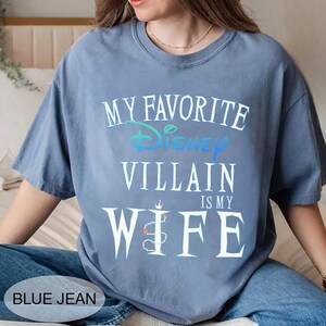 Peut inclure: Un t-shirt bleu avec le texte "MY FAVORITE Disney VILLAIN IS MY WIFE" en blanc et bleu clair. Le mot "Disney" est de couleur sarcelle. Le t-shirt est port&eacute; avec un jean bleu.