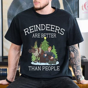 May include: Black t-shirt with the text "REINDEERS ARE BETTER THAN PEOPLE" in white. The graphic features a reindeer, a man playing a lute, and Christmas trees. The shirt is a crew neck.
