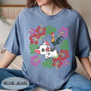 Może przedstawiać: Niebieska koszulka z nadrukiem świni, koguta i kwiat&oacute;w. Świnia jest biała z czarnymi plamami i r&oacute;żowymi uszami. Kogut jest zielony, ż&oacute;łty i czerwony. Kwiaty są czerwone i r&oacute;żowe. Tekst "BLUE JEAN" znajduje się na dole.