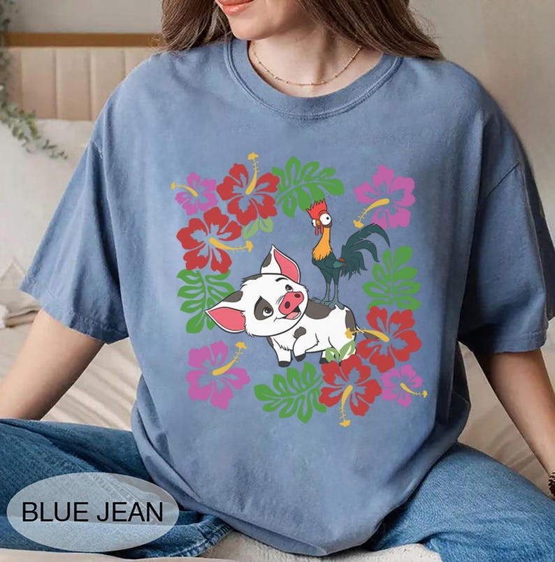 Może przedstawiać: Niebieska koszulka z nadrukiem świni, koguta i kwiat&oacute;w. Świnia jest biała z czarnymi plamami i r&oacute;żowymi uszami. Kogut jest zielony, ż&oacute;łty i czerwony. Kwiaty są czerwone i r&oacute;żowe. Tekst "BLUE JEAN" znajduje się na dole.