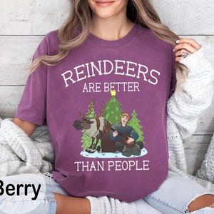 May include: Berry-colored t-shirt with the text "REINDEERS ARE BETTER THAN PEOPLE" in white. The graphic features a reindeer, a man playing a lute, and Christmas trees. The shirt is a crew neck.