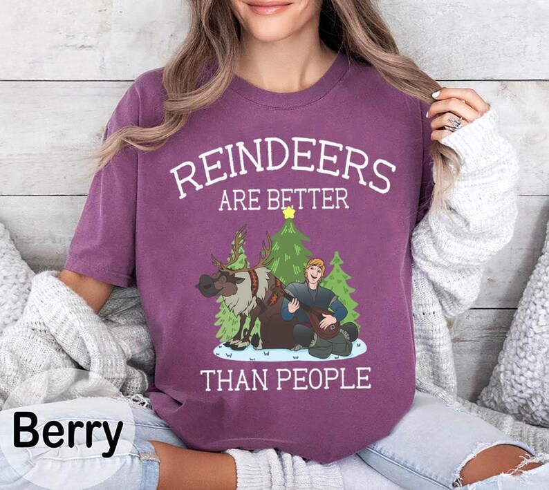 May include: Berry-colored t-shirt with the text "REINDEERS ARE BETTER THAN PEOPLE" in white. The graphic features a reindeer, a man playing a lute, and Christmas trees. The shirt is a crew neck.