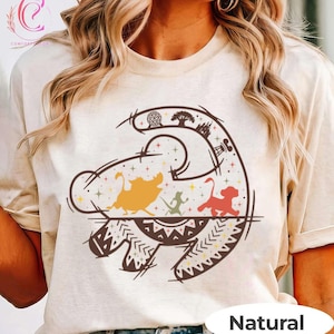 May include: Beige t-shirt featuring a colorful graphic design of a lion's head with various characters and symbols. The word "Natural" is printed at the bottom. The shirt has short sleeves and a crew neck.