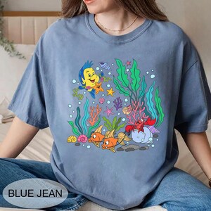 K&ouml;nnte beinhalten: Blaues Jeans-T-Shirt mit einer farbenfrohen Unterwasserszene mit Zeichentrickfischen, Korallen und Blasen. Das Design enth&auml;lt den Text "BLUE JEAN" unten. Das Shirt ist aus weichem Material.