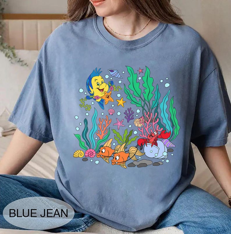 K&ouml;nnte beinhalten: Blaues Jeans-T-Shirt mit einer farbenfrohen Unterwasserszene mit Zeichentrickfischen, Korallen und Blasen. Das Design enth&auml;lt den Text "BLUE JEAN" unten. Das Shirt ist aus weichem Material.
