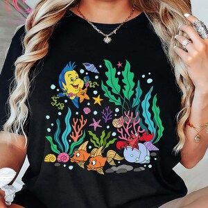 K&ouml;nnte beinhalten: Schwarzes T-Shirt mit einer farbenfrohen Cartoon-Illustration einer Unterwasserszene. Das Design umfasst einen gelben Fisch, Seesterne, orangefarbene Fische, eine rote Krabbe sowie verschiedene Wasserpflanzen und Korallen. Das Design ist vor einem Hintergrund aus Blasen und Felsen.