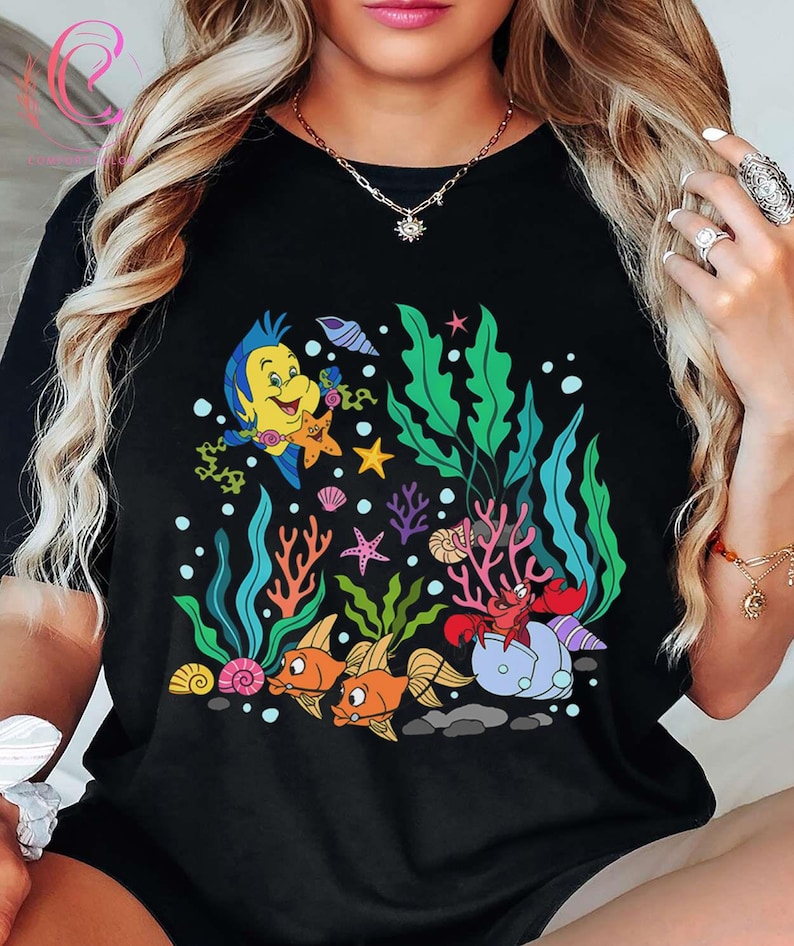 K&ouml;nnte beinhalten: Schwarzes T-Shirt mit einer farbenfrohen Cartoon-Illustration einer Unterwasserszene. Das Design umfasst einen gelben Fisch, Seesterne, orangefarbene Fische, eine rote Krabbe sowie verschiedene Wasserpflanzen und Korallen. Das Design ist vor einem Hintergrund aus Blasen und Felsen.