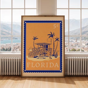 A framed orange and blue Florida beach poster stands on a wooden floor in front of a large window overlooking a mountainous landscape.