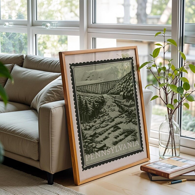 A large framed print, designed like a vintage postage stamp, depicts a river valley with a viaduct, trees, and buildings, on a wooden floor next to a couch.