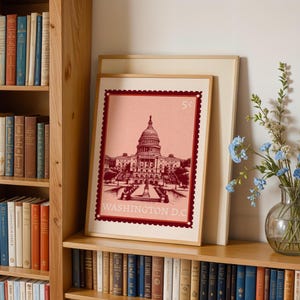 Puede incluir: Impresión enmarcada del edificio del Capitolio de los Estados Unidos, diseñada para parecerse a un sello postal. La impresión es en tonos de rojo y rosa, con el texto "WASHINGTON D.C." en la parte inferior y "5¢" en la parte superior. La impresión se exhibe en una estantería de madera con libros.