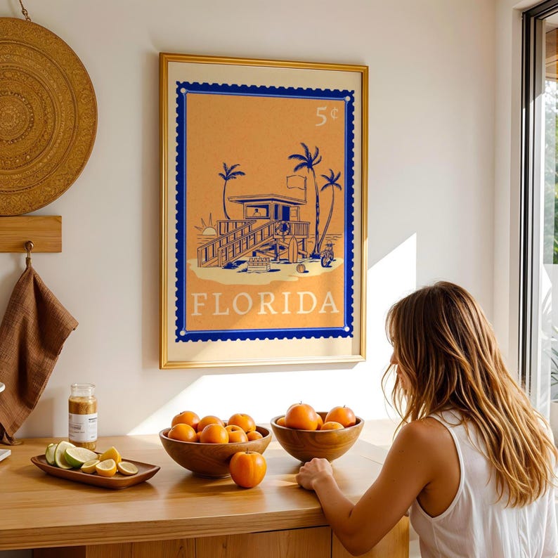 A framed orange and blue Florida beach poster hangs on a white wall above a wooden counter with bowls of oranges and a woman looking at them.