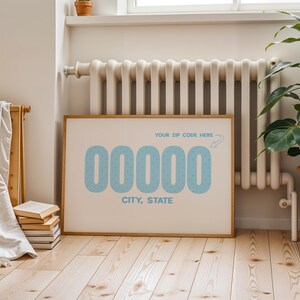 May include: A framed print with a light beige background features the text "YOUR ZIP CODE HERE" and a series of light blue numbers and the words "CITY, STATE". The print is in a wooden frame and is displayed against a white wall.