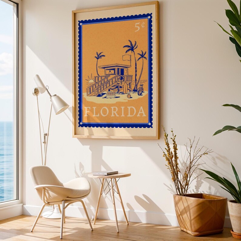A framed orange and blue Florida beach poster hangs on a beige wall in a modern room with a white chair and a large window.