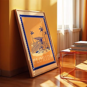 A framed orange and blue Florida beach poster leans against a yellow wall on a wooden floor, with a clear orange side table nearby.