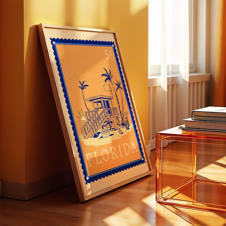 A framed orange and blue Florida beach poster leans against a yellow wall on a wooden floor, with a clear orange side table nearby.