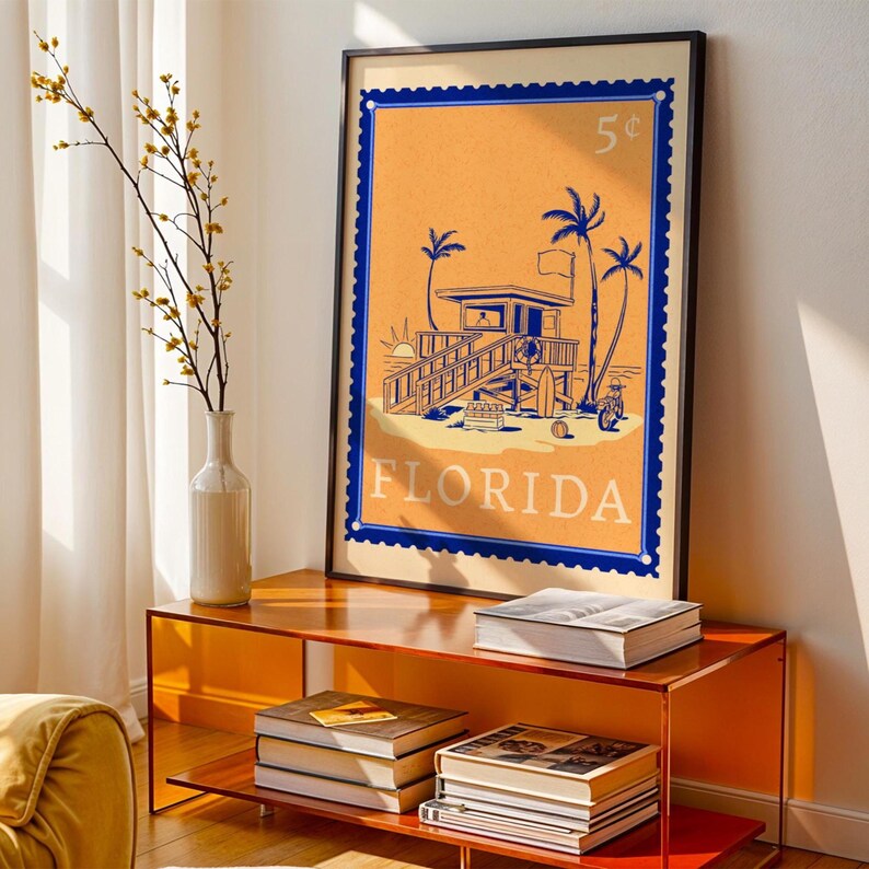 A framed orange and blue Florida beach poster stands on an orange console table next to a white vase with yellow flowers.