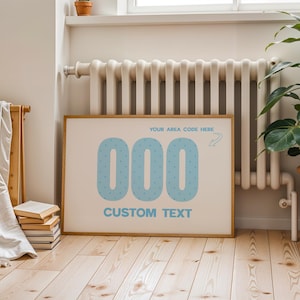 May include: A rectangular framed print featuring the light blue numbers 000 with small dots, the text YOUR AREA CODE HERE with an arrow pointing to the numbers, and the text CUSTOM TEXT in light blue on a cream background.