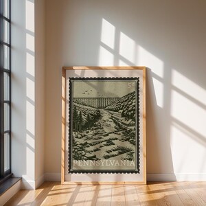 A large framed print, designed like a vintage postage stamp, depicts a river valley with a viaduct, trees, and buildings, standing on a wooden floor against a wall.