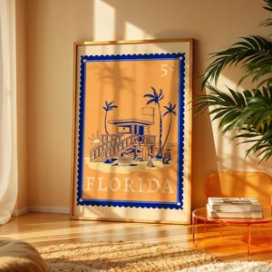 A large framed orange and blue Florida beach poster leans against a yellow wall next to a green plant and a clear orange side table.