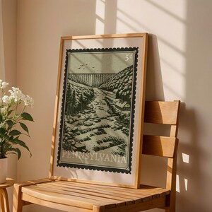 A framed print, designed like a vintage postage stamp, depicts a river valley with a viaduct, trees, and buildings, leaning against a wooden chair next to a plant.