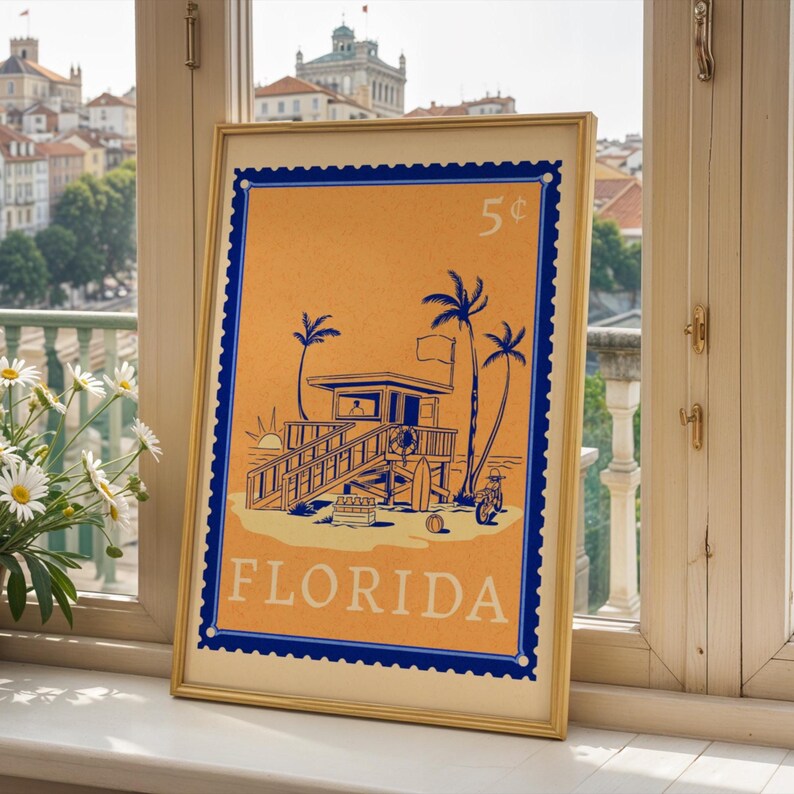 A framed orange and blue Florida beach poster sits on a white windowsill overlooking a city with a large building in the background.