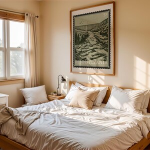 A large framed print, designed like a vintage postage stamp, depicts a river valley with a viaduct, trees, and buildings, hanging on a wall above a bed.