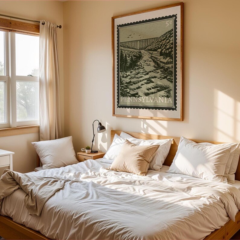 A large framed print, designed like a vintage postage stamp, depicts a river valley with a viaduct, trees, and buildings, hanging on a wall above a bed.