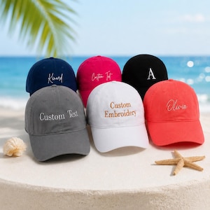 Op de afbeelding: Een verzameling van zes baseballpetten in diverse kleuren, waaronder marineblauw, roze, zwart, grijs, wit en koraal. Elke pet heeft een aangepaste borduursel met tekst of een enkele letter. De petten worden getoond op een zanderige ondergrond met een strandachtergrond.
