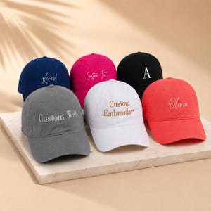 May include: Six baseball caps in a range of colours: navy, pink, black, grey, white, and red. Each cap has custom embroidery or text, such as names or phrases, in a contrasting colour. The caps are displayed on a neutral surface.