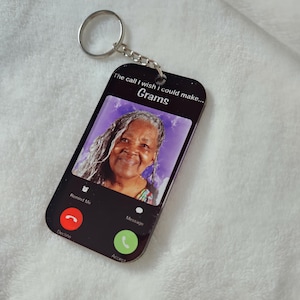 May include: Black phone-shaped keychain with a photo of an older woman with gray hair. The text on the keychain reads "The call I wish I could make... Grams".