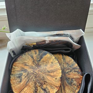May include: Two round, wooden coasters with a black, gold, and brown marbled design. The coasters are in a black box with a black satin lining.