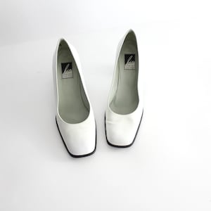 May include: A pair of white leather pumps with a square toe and a low heel. The shoes have a black label on the inside that says "Liz Claiborne New York".
