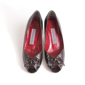 Pode incluir: Um par de sapatos de salto alto peep-toe marrons com um detalhe de laço. Os sapatos são feitos de couro texturizado e têm um forro vermelho. O nome da marca "Evan Picone" está impresso no interior dos sapatos.