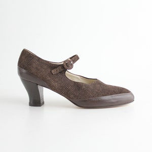 May include: Brown Mary Jane style shoe with a strap and buckle closure. The shoe has a textured fabric upper and a dark brown heel. The heel is approximately 3 inches high.