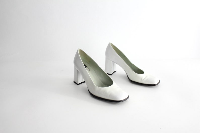 Puede incluir: Un par de zapatos de sal&oacute;n de cuero blanco con tac&oacute;n de bloque. Los zapatos tienen una puntera cuadrada y un dise&ntilde;o sencillo.