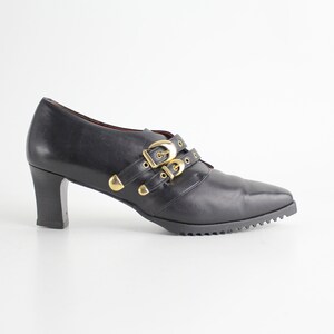 May include: Black leather heeled shoes with gold buckle accents.