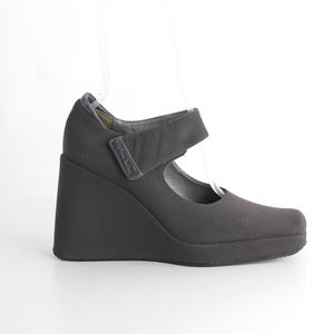May include: Black wedge heel shoe with a strap across the top of the foot. The shoe has a rounded toe and a thick, angled heel. The shoe is made of a matte material and sits on a white surface.